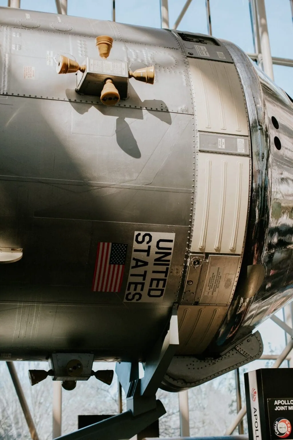 Riveted metal spacecraft section marked UNITED STATES with an American flag decal, on display in a museum hall