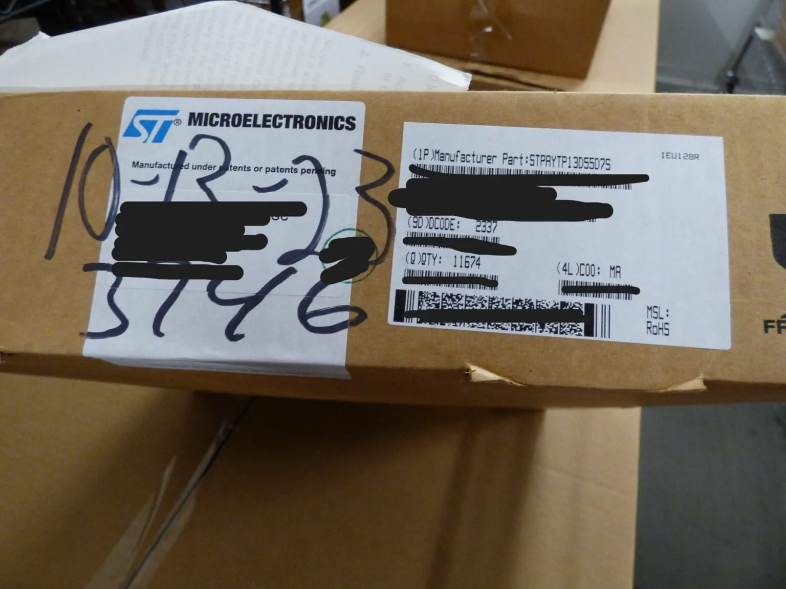 Cardboard box with an ST Microelectronics label, a part label showing quantity 11674 and a handwritten date, some text blacked out