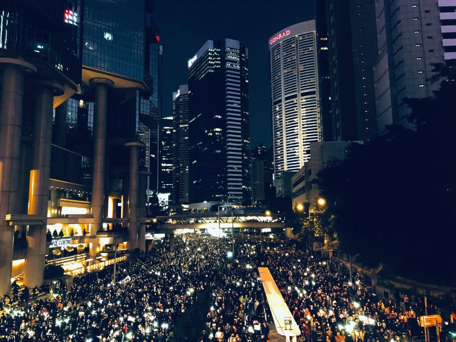 Night view of a dense crowd lit by phone lights filling a street between skyscrapers, one tower marked CONRAD