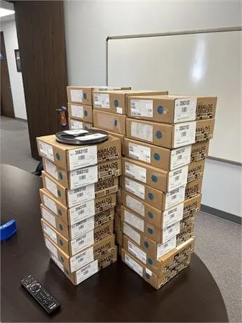 Tall stacks of brown boxes labeled Analog Devices on a conference table with a reel on top and a remote in front