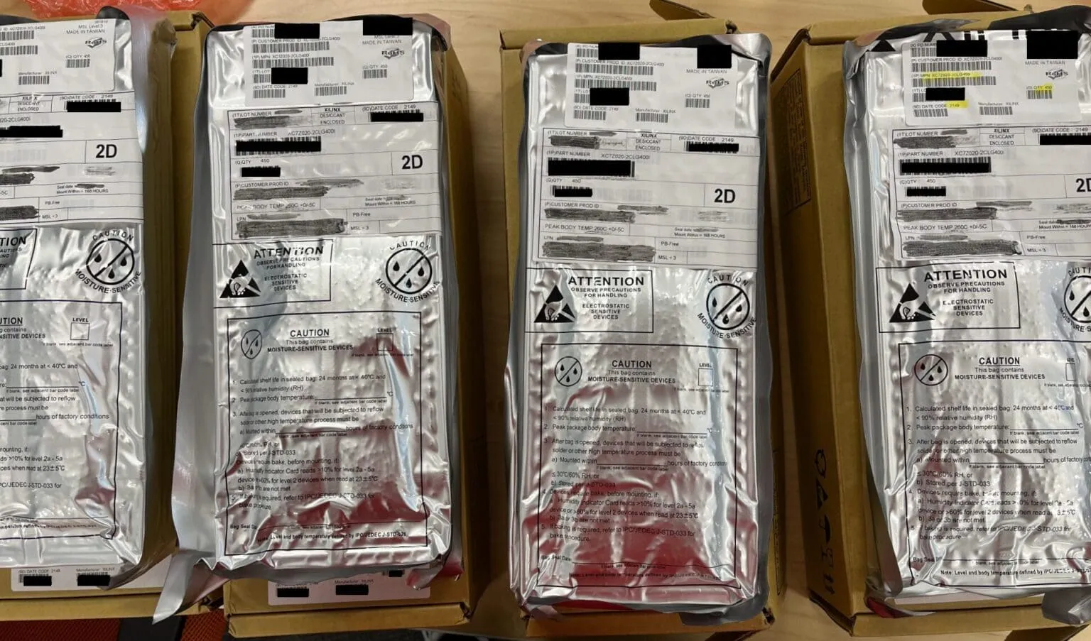 Four silver moisture-barrier bags on cardboard boxes with Xilinx labels and ESD and moisture warnings, some text blacked out