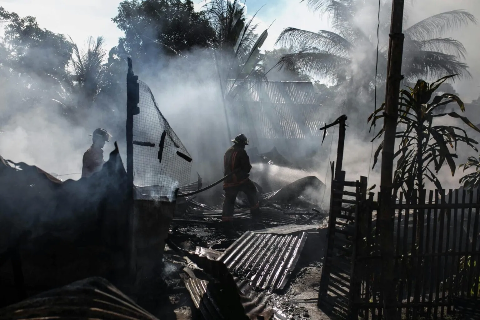 Two firefighters in helmets in heavy smoke spraying a hose over burned corrugated metal debris near palm trees
