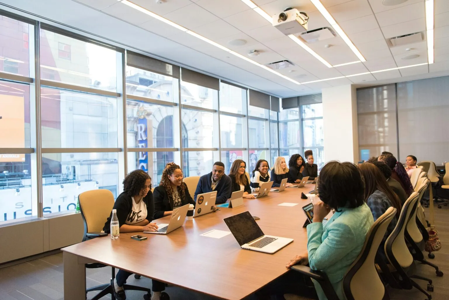People with laptops seated along a long wooden conference table in a bright room with large windows