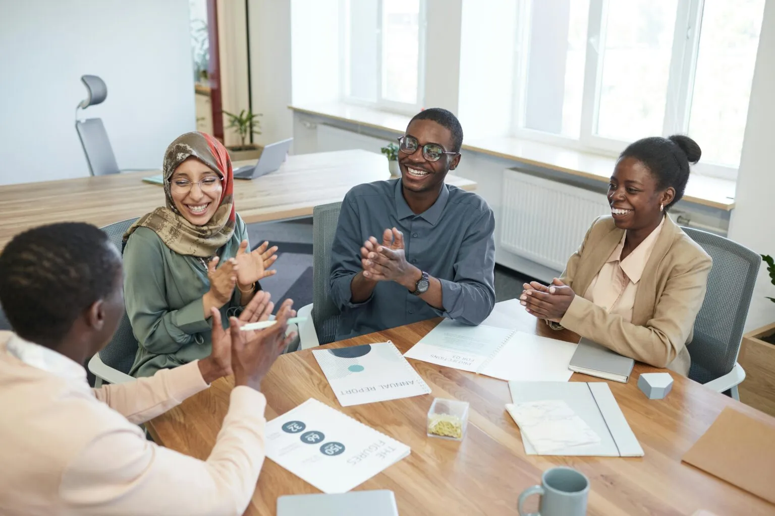 Four smiling colleagues applauding around a wooden meeting table with annual report and figures handouts