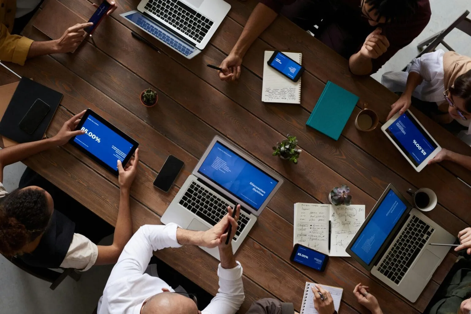 Overhead view of people around a wooden table with laptops, tablets and notebooks showing blue slides