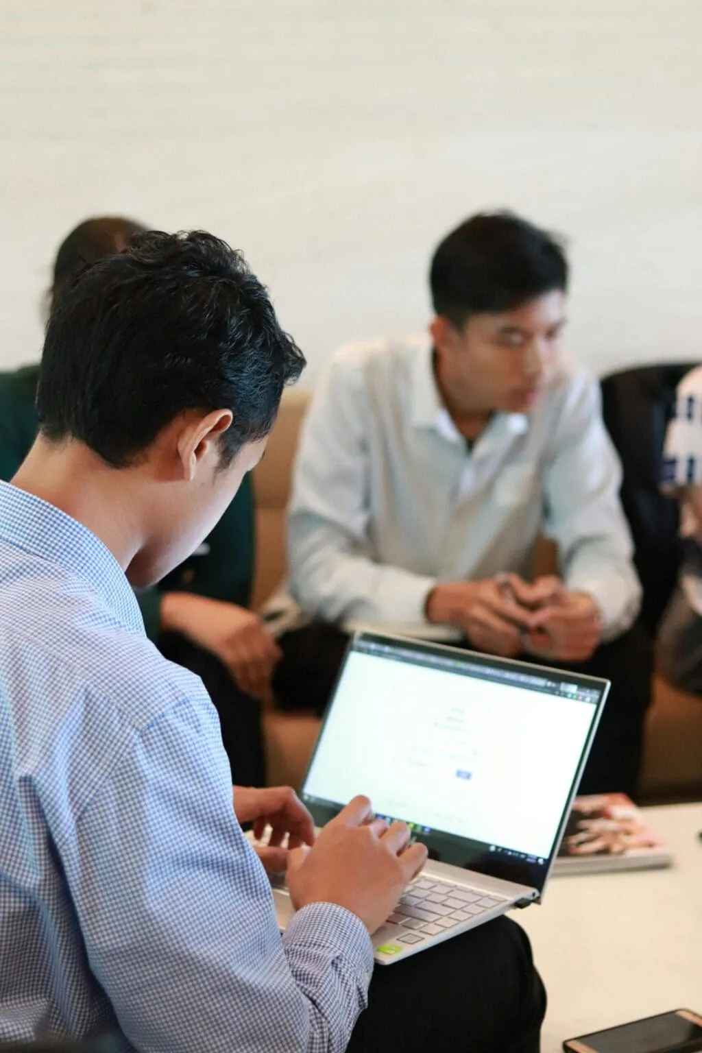 Man in a checked shirt typing on a laptop in the foreground while two other men talk on a sofa behind him