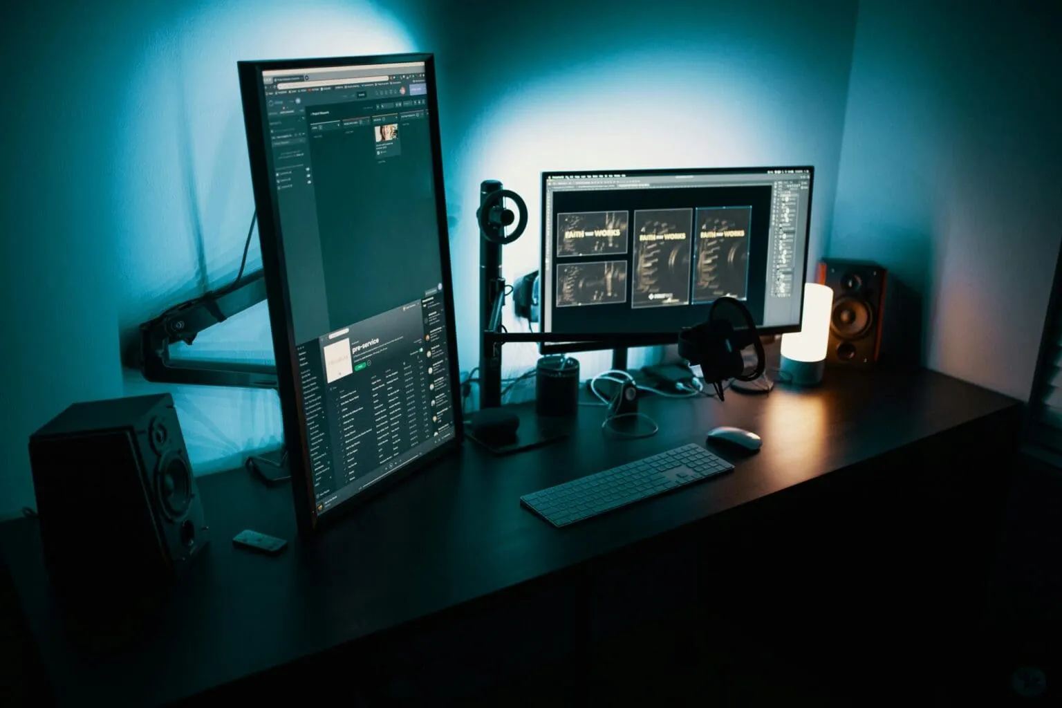 Dark desk lit in blue, with a vertical monitor, a wide monitor showing design work, a keyboard, mouse, headphones and two speakers