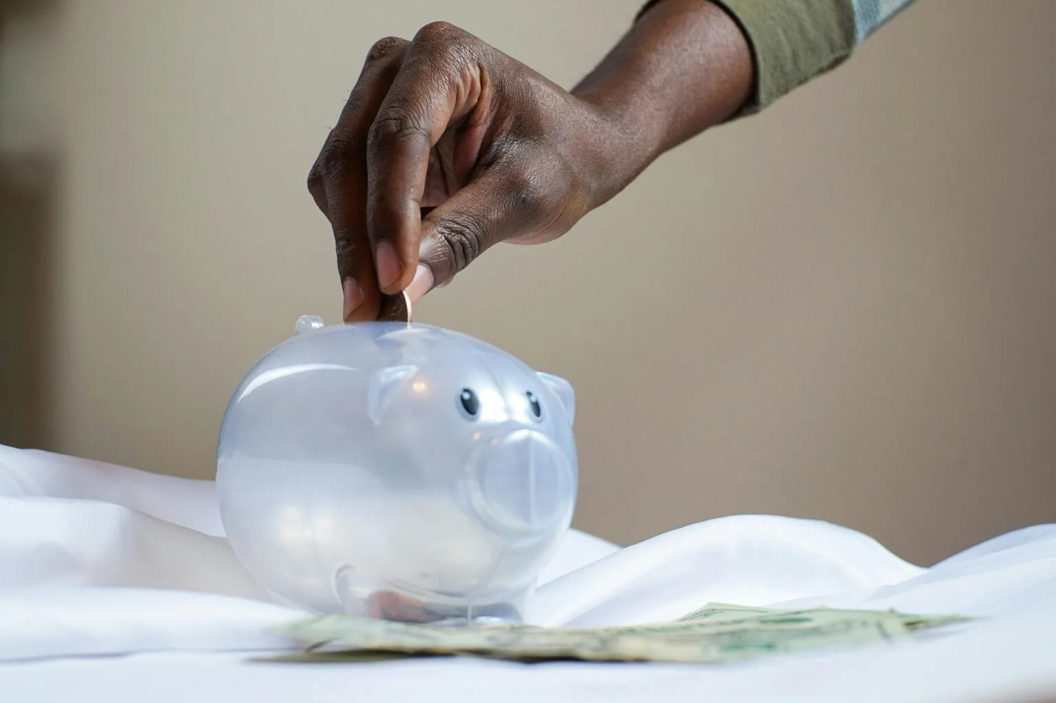 Hand dropping a coin into a clear plastic piggy bank, with banknotes lying on white fabric in front