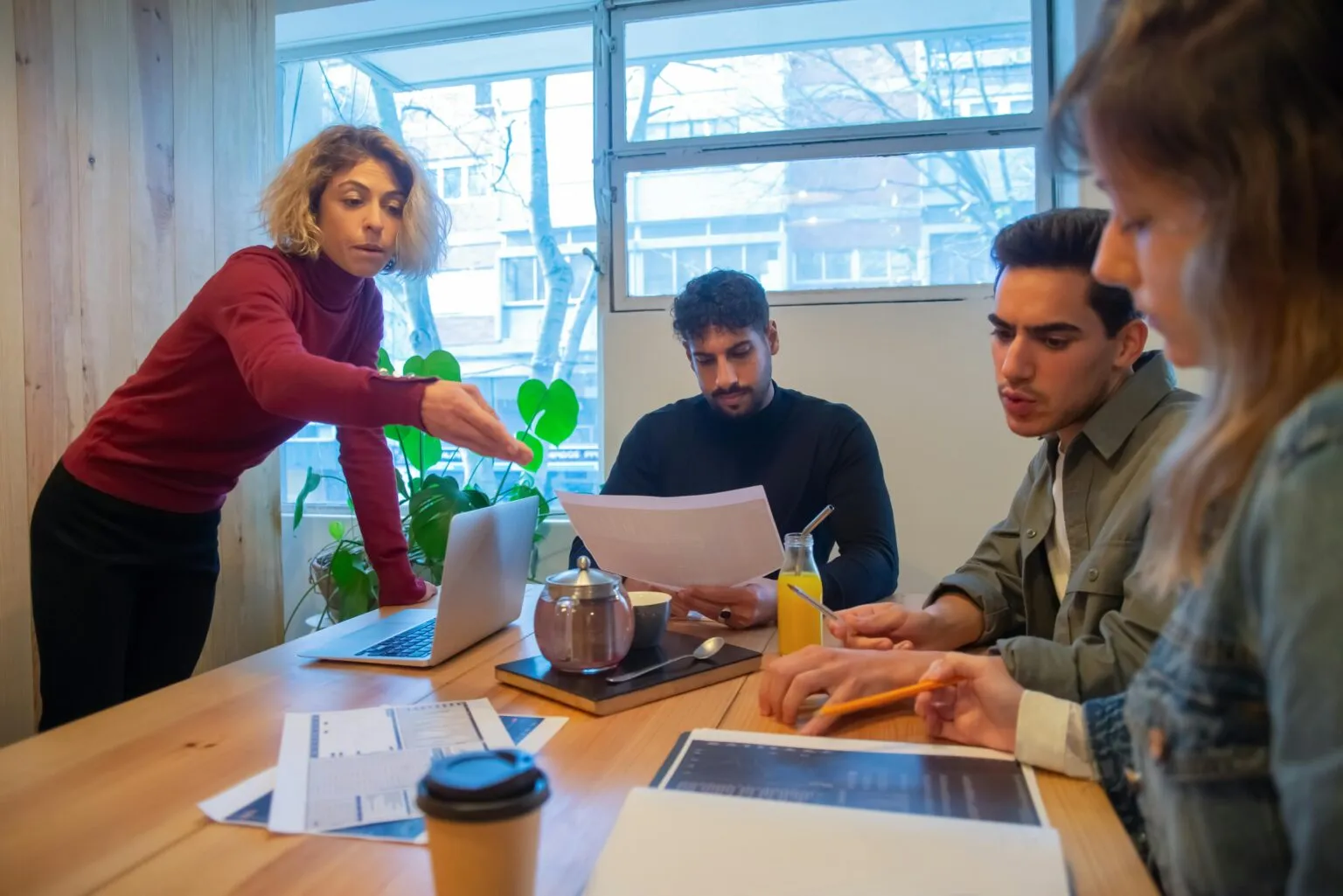 Four people around a wooden table with papers and a laptop while a standing woman points toward the screen