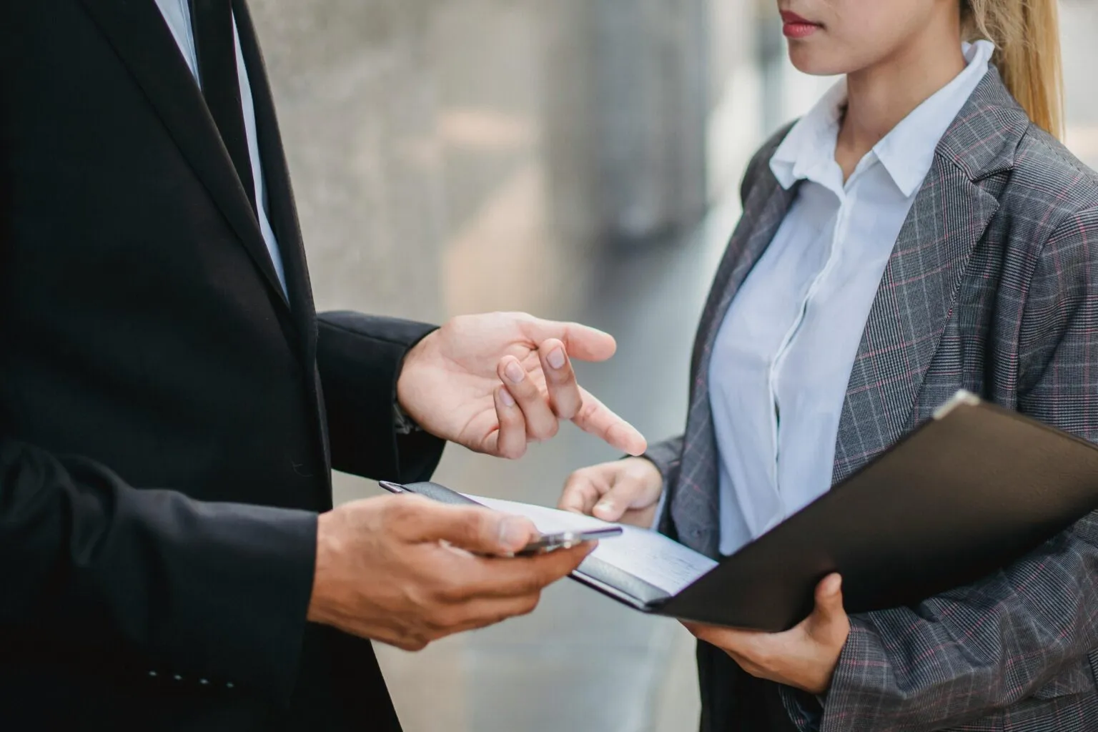 Cropped view of two people in business attire, one gesturing while holding a phone, the other holding a folder