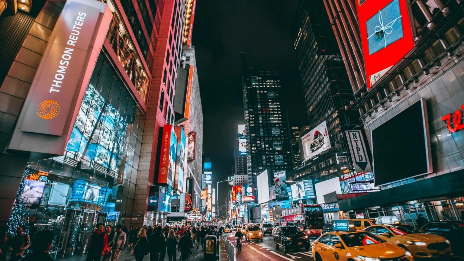 Night street lined with lit billboards and glass towers, a crowd on the sidewalk and yellow taxis on the road
