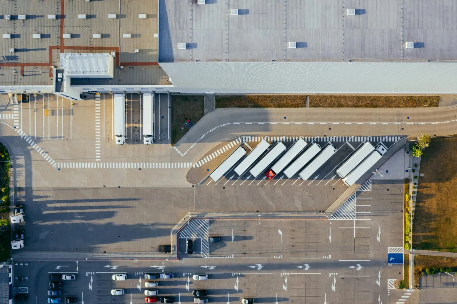 Aerial top-down view of a large warehouse roof, trucks parked at loading bays, and a car park below