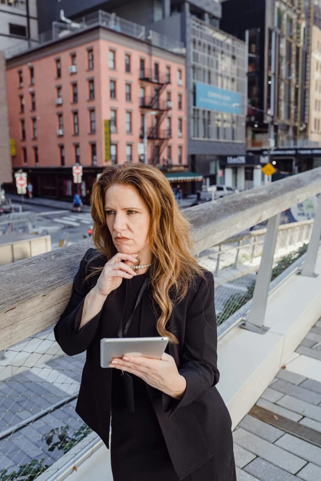Woman in a black blazer holding a tablet and resting her chin on her hand on an elevated walkway above a city street