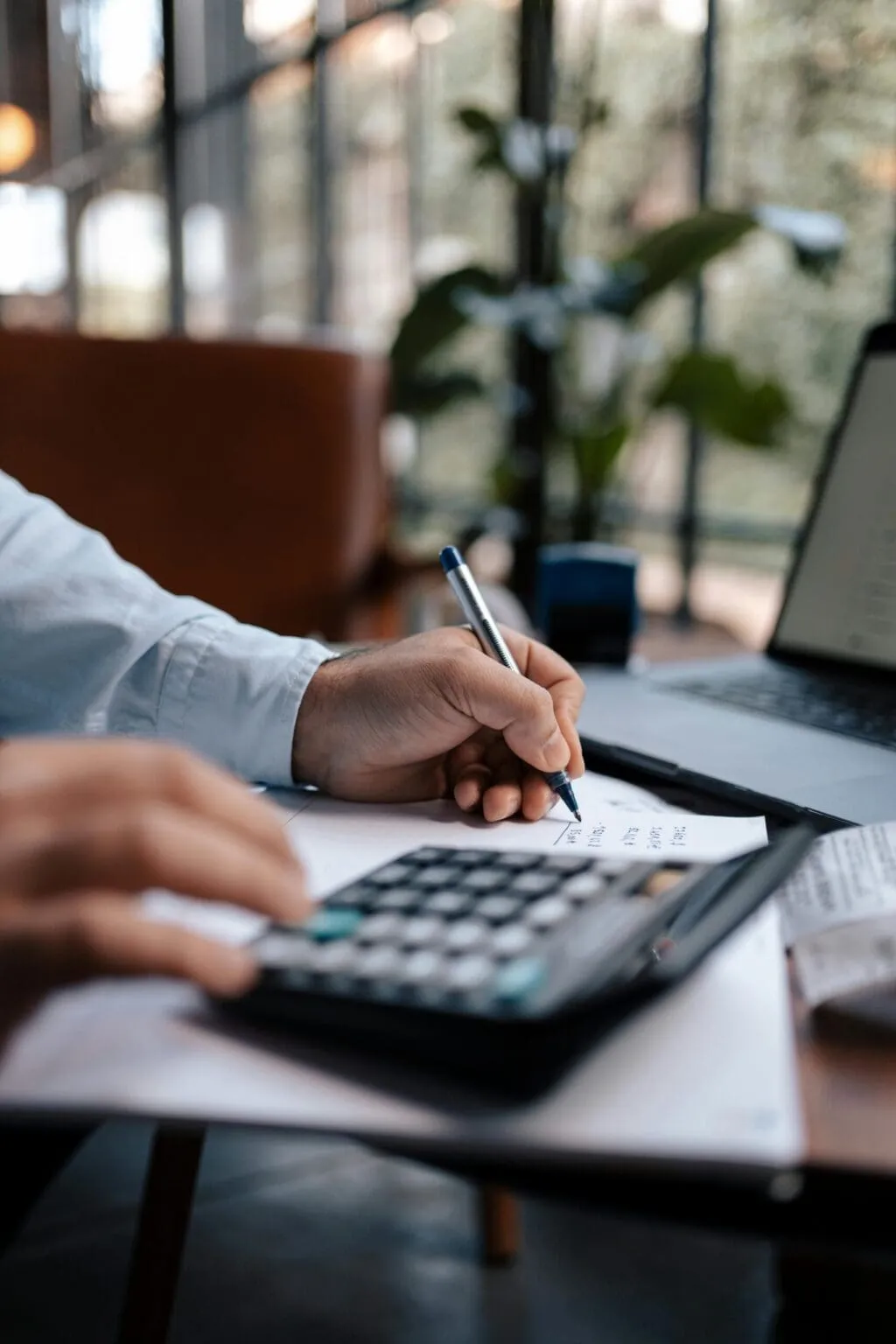 Person writing on paper with a pen while using a calculator at a desk, a laptop and houseplant in the background