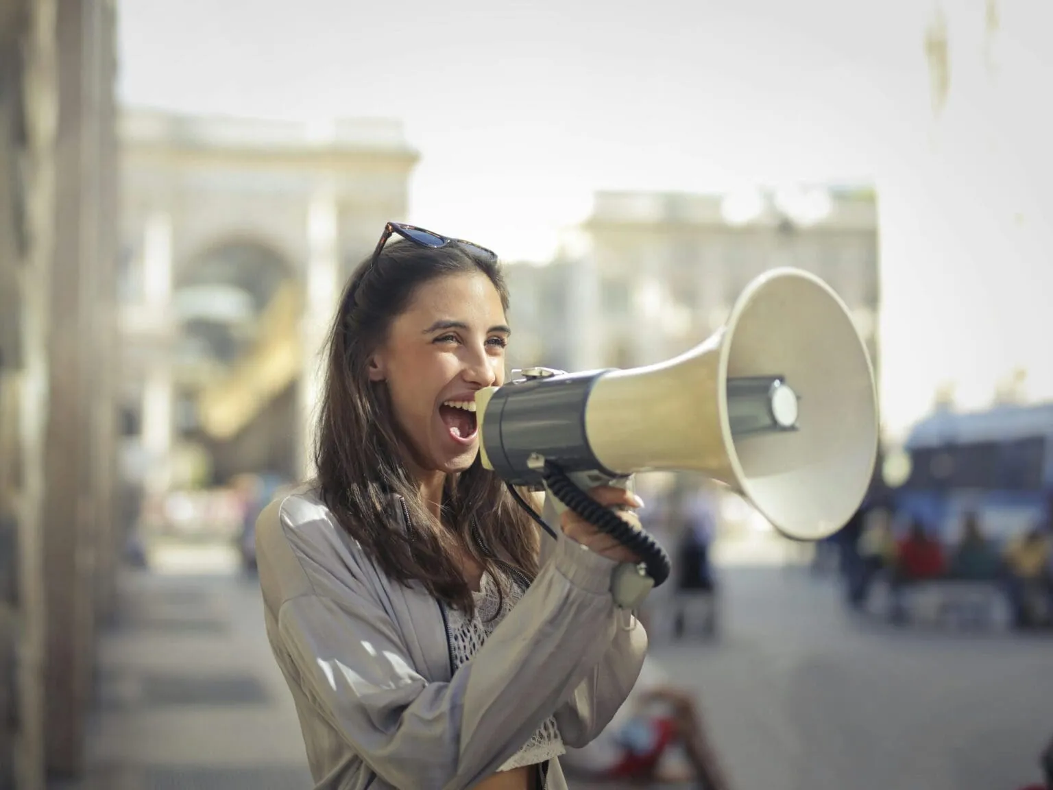 Smiling woman with sunglasses on her head shouting into a megaphone on a blurred city street
