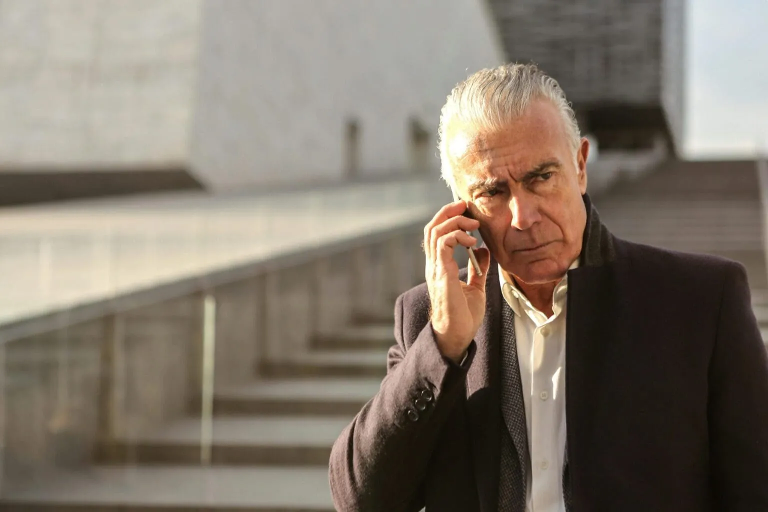 Gray-haired man in a dark coat and white shirt frowning as he talks on a phone in front of concrete steps