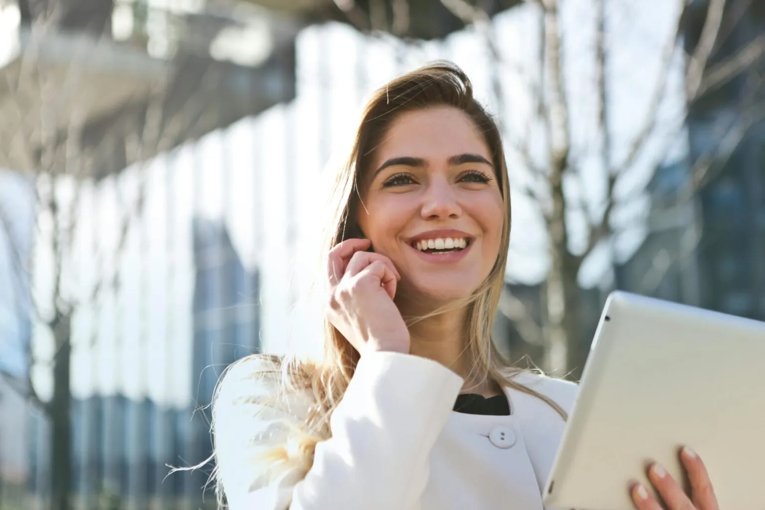 Smiling woman in a white coat holding a phone to her ear and carrying a tablet, with leafless trees and glass buildings blurred behind her