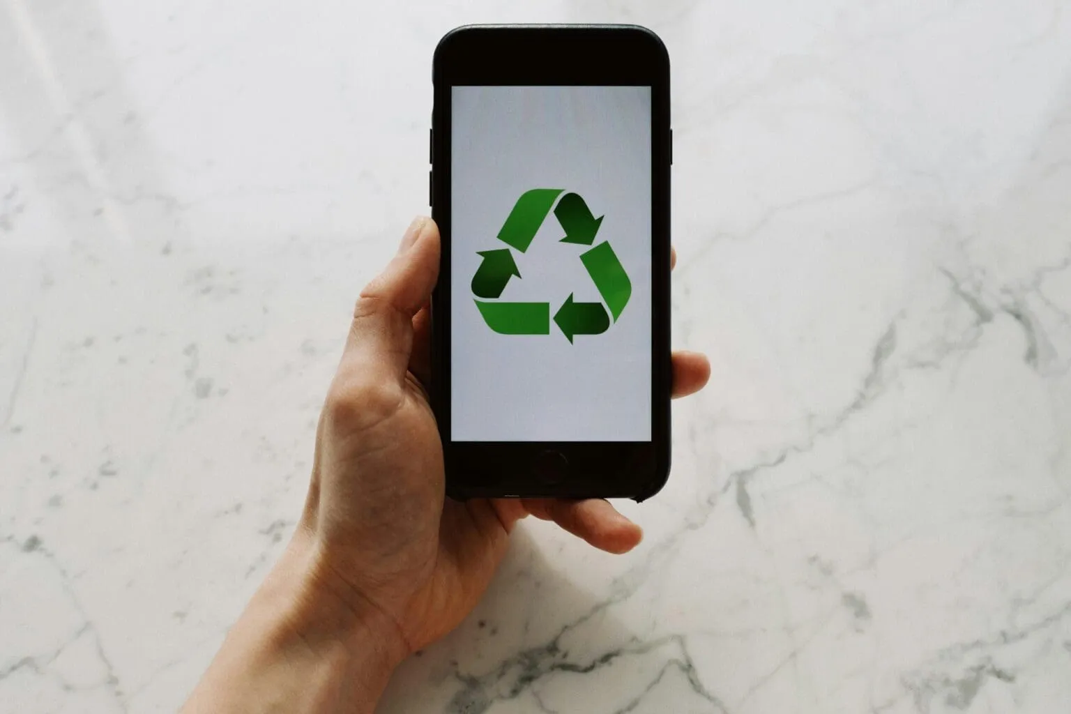 Hand holding a smartphone whose screen shows a green three-arrow recycling symbol, against a white marble surface