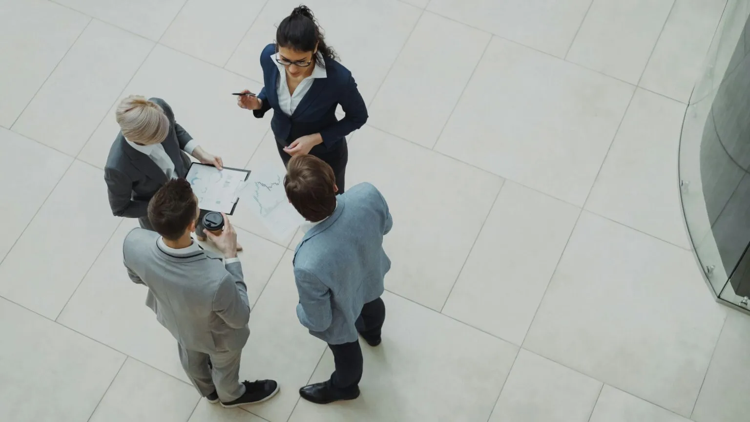 Overhead view of four people in business attire standing in a circle on a tiled floor, reviewing papers on a clipboard