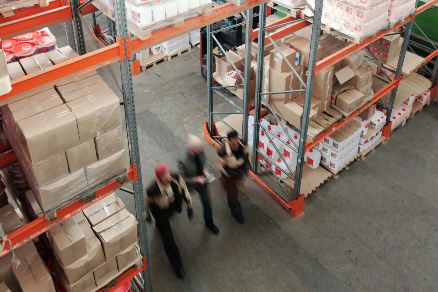 Overhead view of three blurred workers walking past orange warehouse racking stacked with boxes and white jugs