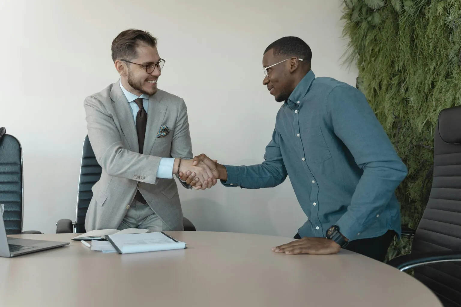 Two men shaking hands across a round table with a laptop and papers, a green plant wall behind them