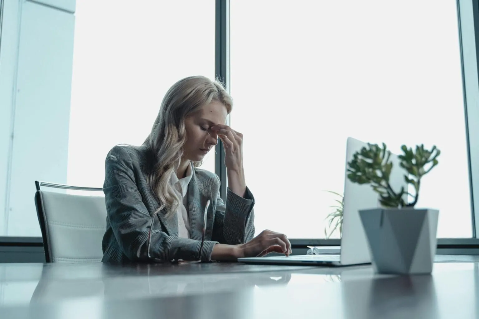Woman in a gray blazer pinching the bridge of her nose at a desk with a laptop and a potted plant
