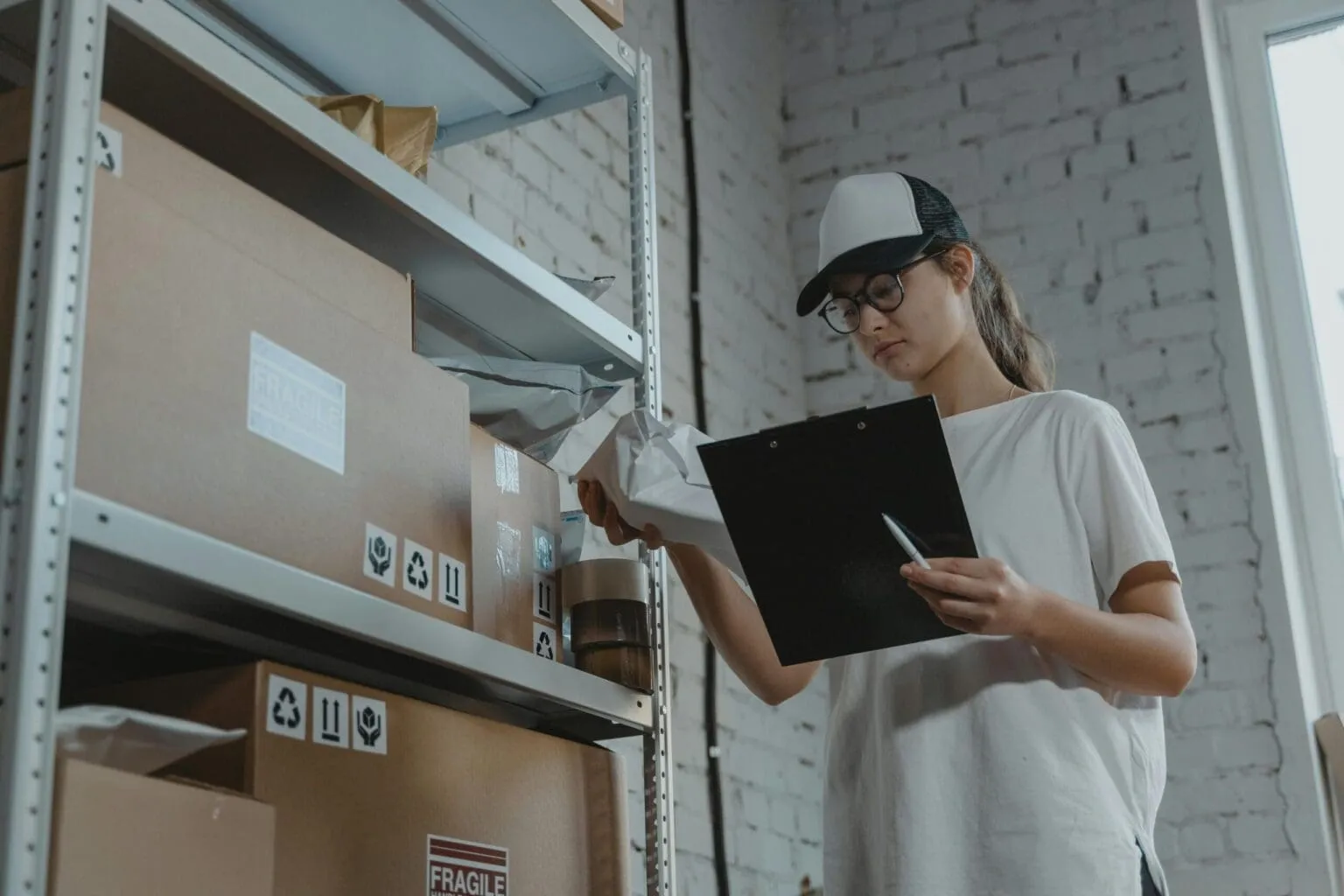Woman in a cap and glasses holding a clipboard beside metal shelves of cardboard boxes marked FRAGILE