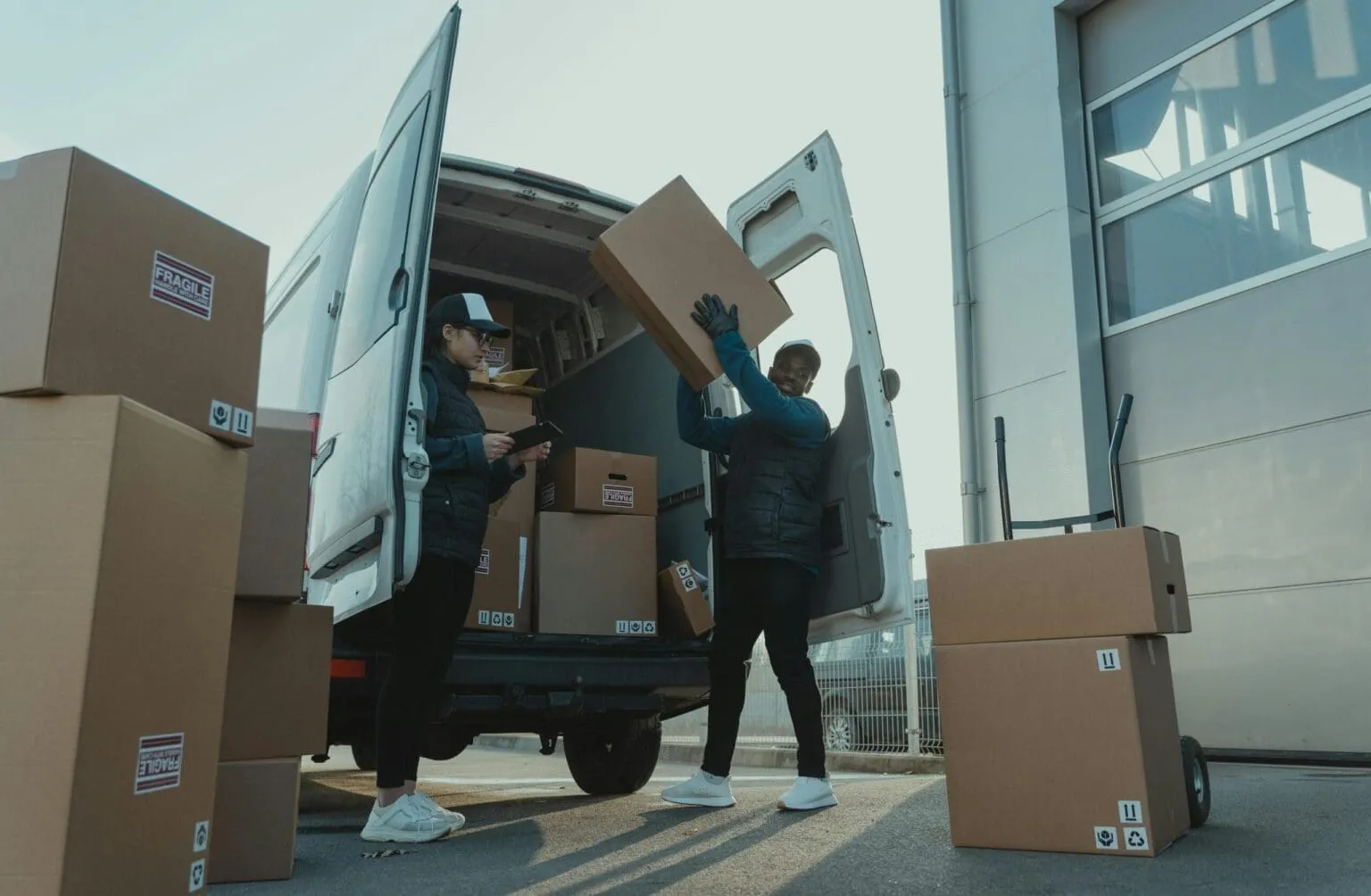 Two people unloading cardboard boxes from a van with open rear doors, more boxes stacked beside it, one marked Fragile