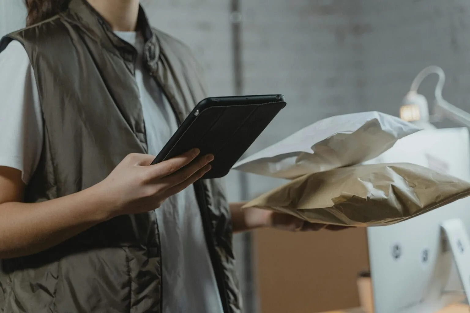 Person in an olive vest holding a black tablet in one hand and two padded shipping envelopes in the other, with boxes behind