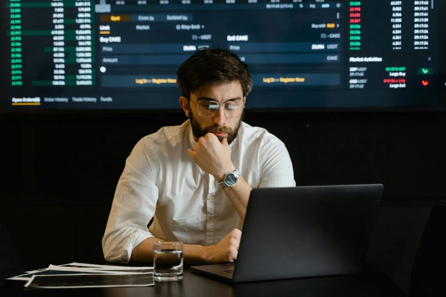 Bearded man with glasses resting his chin on his fist, looking at a laptop in front of a wall screen of trading data