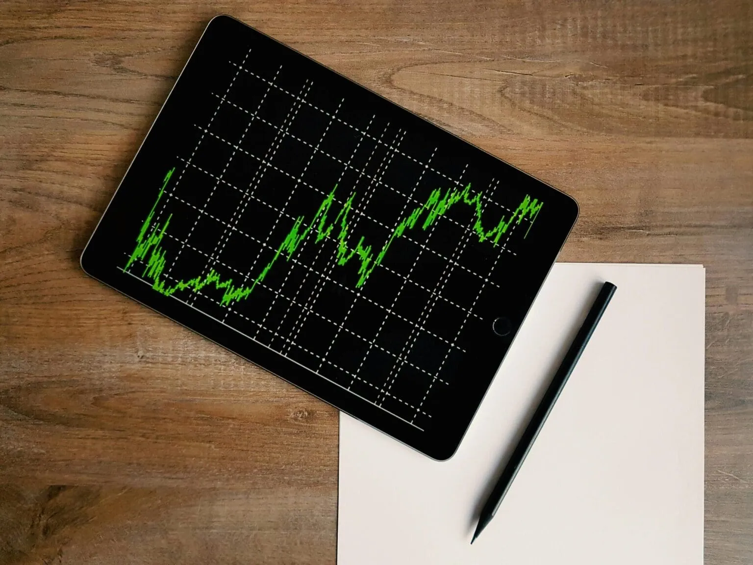 Tablet showing a green fluctuating line chart on a dark grid, beside a blank sheet of paper and a black pencil on a wooden desk