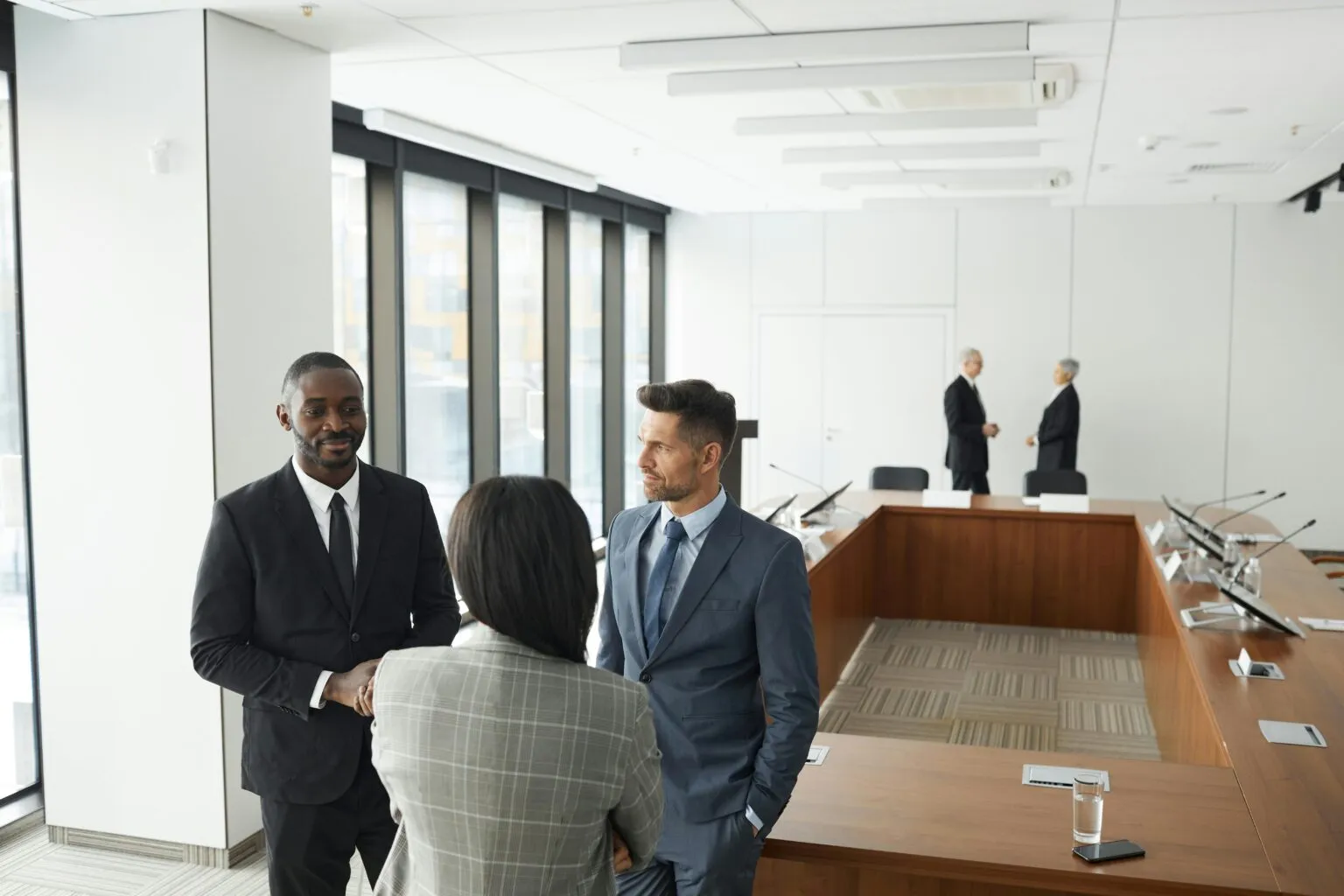 Three people in business suits talking beside a long wooden conference table, with two more people standing in the background