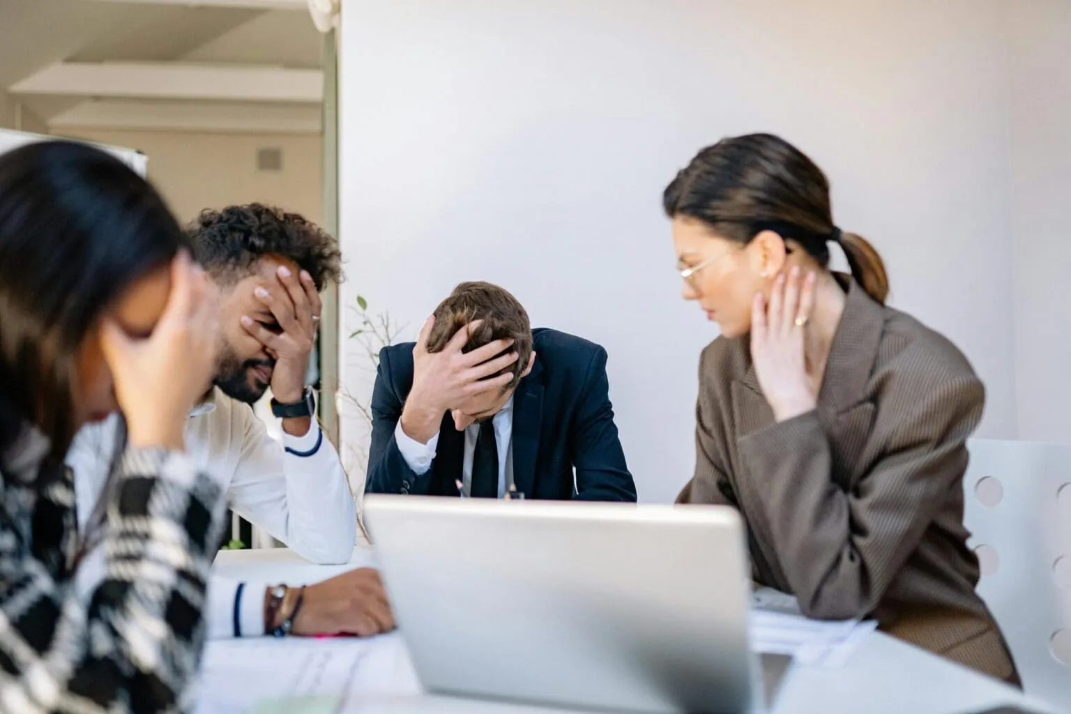 Four colleagues at a meeting table, three with a hand on their head looking stressed, with a laptop in the foreground